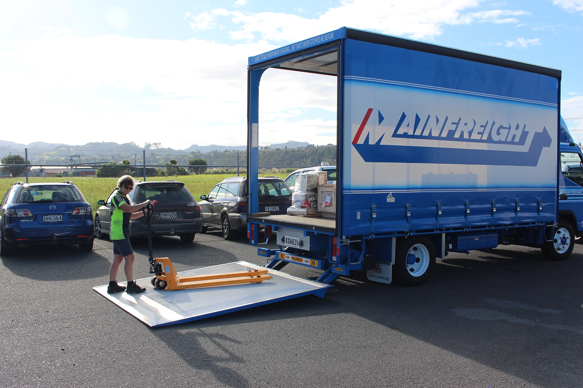 A Mainfreight truck with its Palfinger tail lift lowered, as a worker in a high-visibility shirt uses a pallet jack to move goods onto the lift in a parking area with cars and a scenic rural background.